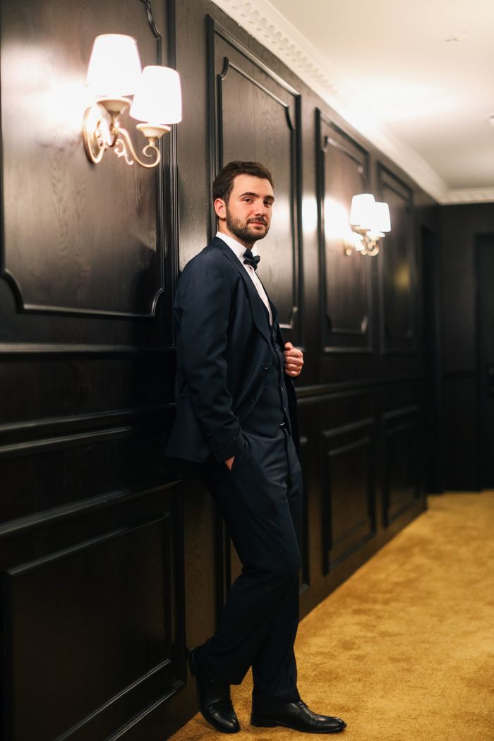 Stylish man in a tuxedo leaning against dark wooden paneling in a classy hallway.