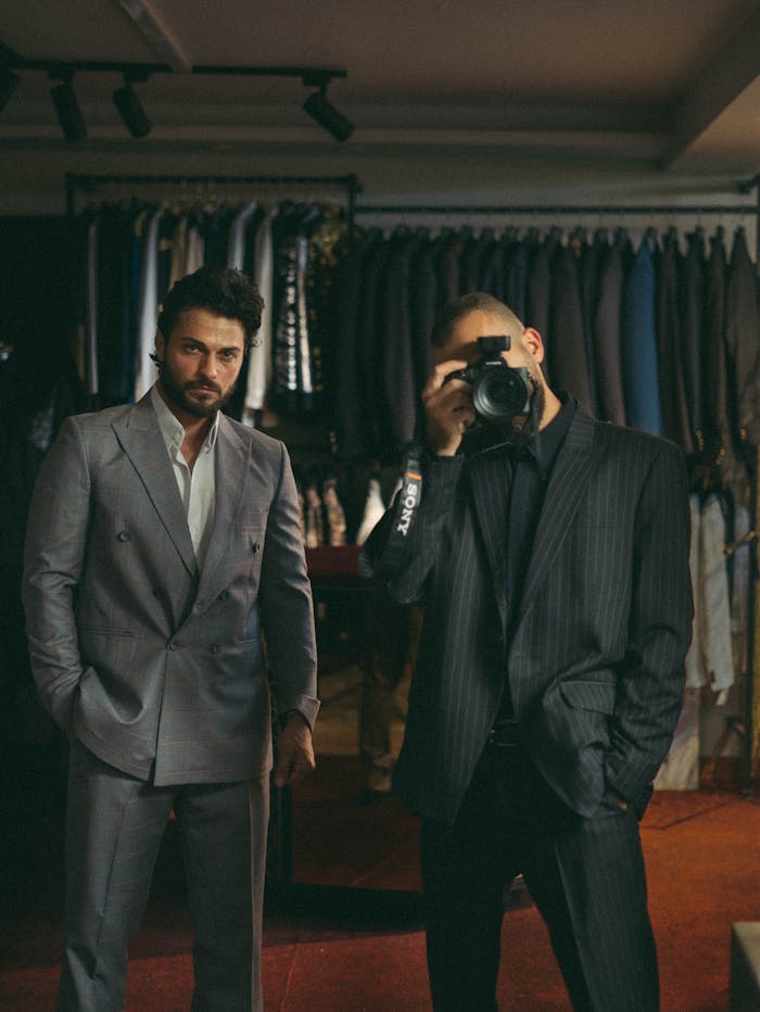 about-us-01 Two men in suits, one taking a photo, in a fashion boutique setting.